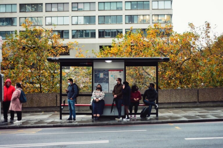 A group of people wait at a bus stop under a canopy, with autumn trees and a modern building in the background.
