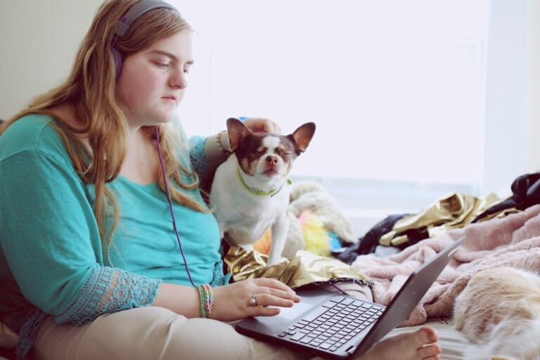 A young woman with blonde hair wearing headphones and a teal shirt sits on a bed with a laptop and two dogs.