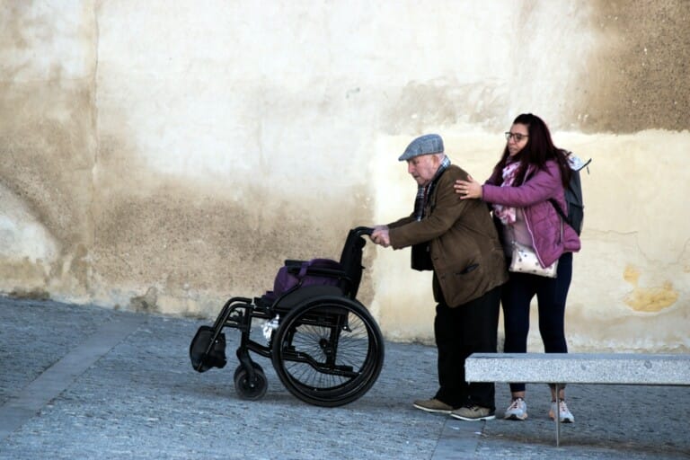An elderly man in a flat cap and brown jacket pushes a wheelchair while a younger woman in a purple jacket walks beside him, placing her hand on his shoulder.