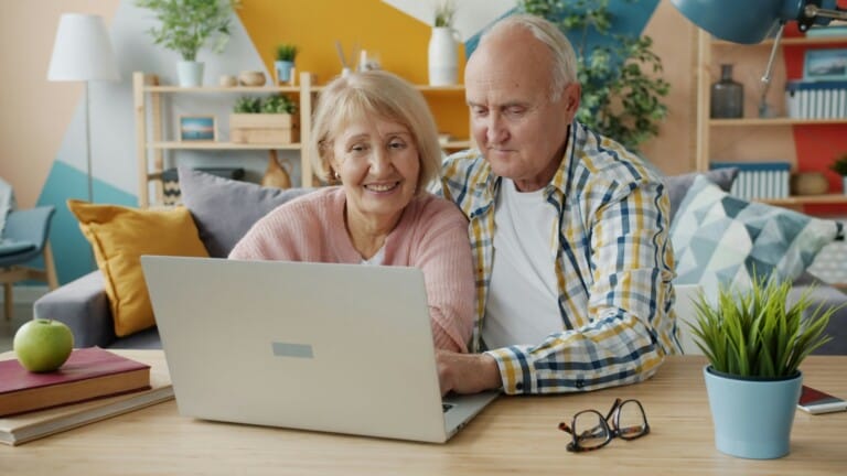An elderly couple smiles while looking at a laptop on a wooden table.